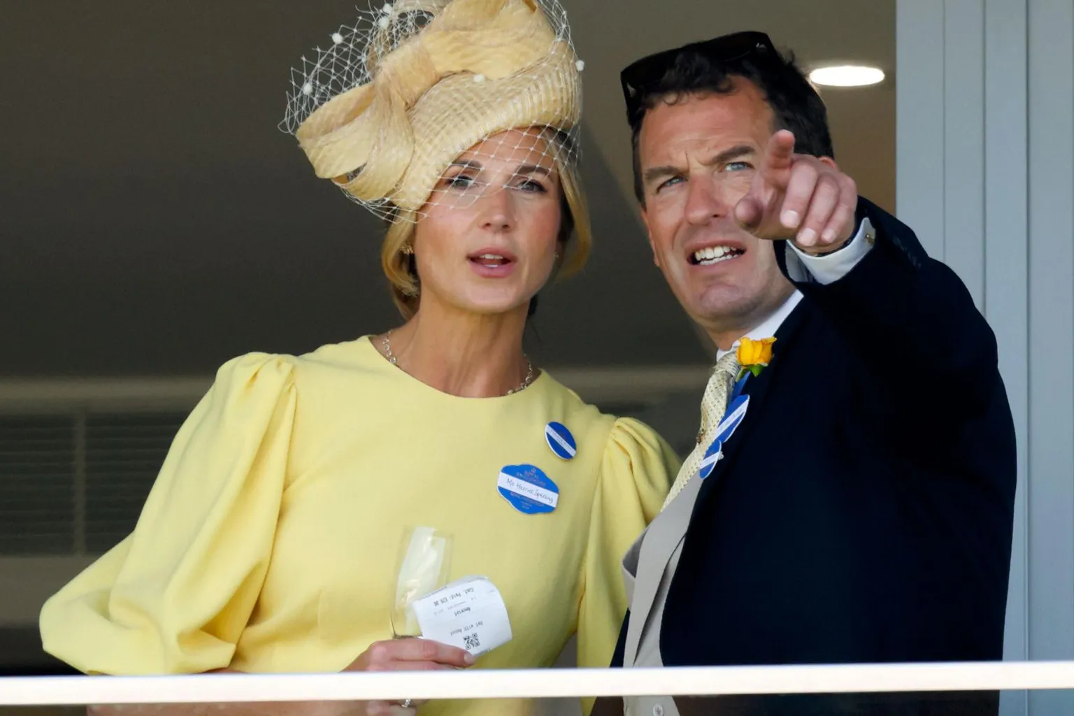 Max Mumby/Indigo/ Harriet Sperling and Peter Phillips watch the racing at Ladies Day at Royal Ascot. She is wearing a yellow dress and hat and he is wearing a yellow flower and is pointing into the distance