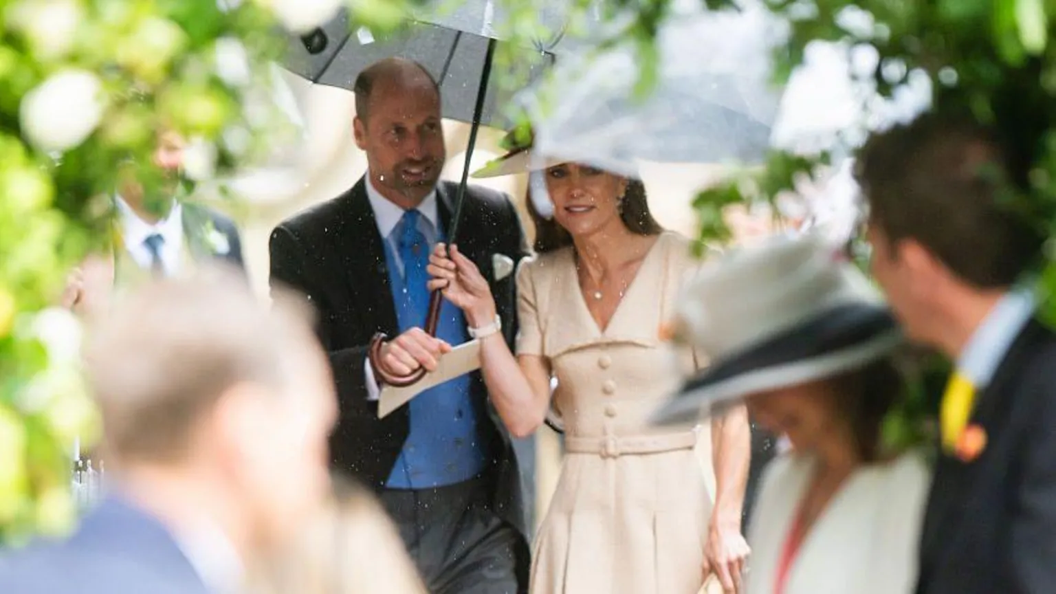 Samir Hussein/WireImage via Prince William holds an umbrella over Kate Middleton as they arrive at the wedding. Kate is wearing a cream dress, while William is wearing a suit with blue waistcoat