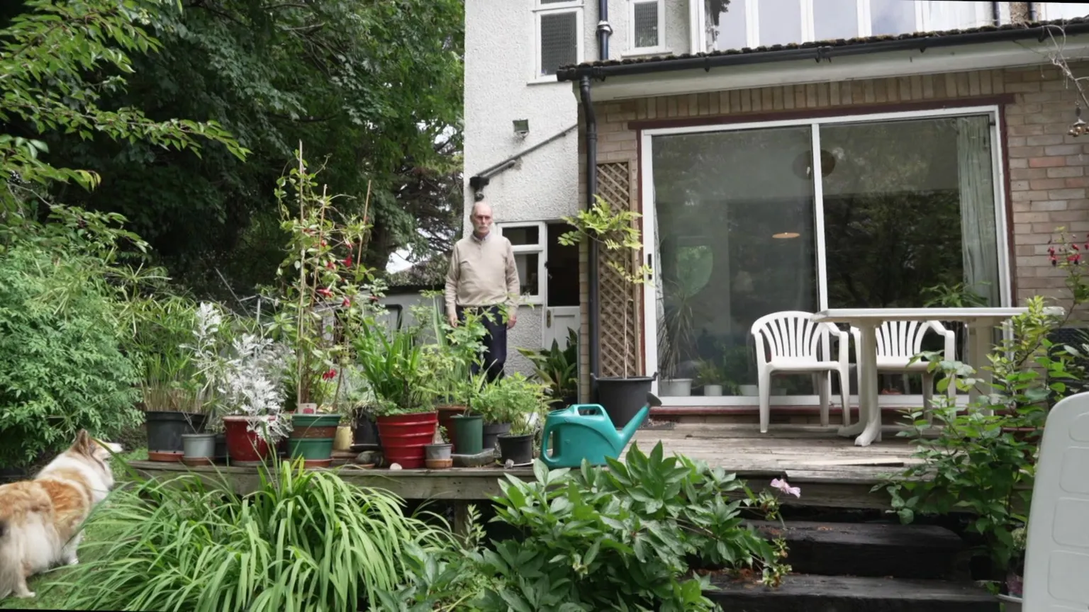 Jim in a beige jumper stands on a wooden decking area outside a house, surrounded by potted plants and greenery, with a dog in the foreground