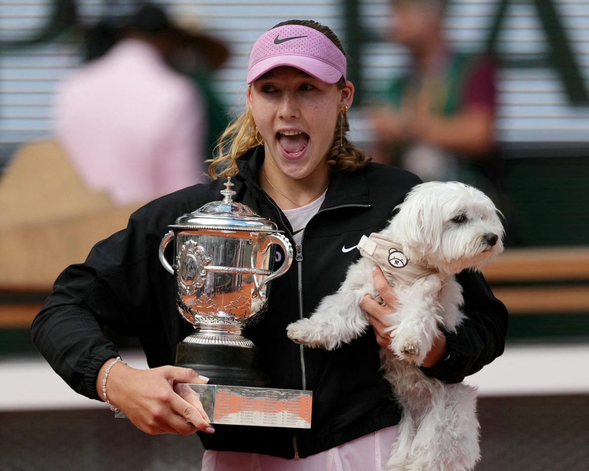 Tennis player Mirra Andreeva, wearing a pink visor and a black zipped jacket over a light pink tennis dress, holds the French Open women's trophy in one hand and a dog in the other.