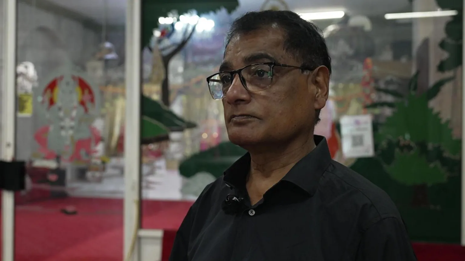Dr Satyavir Singhal wears glasses and has a black shirt on. A mic is attached to his collar. Glass doors are behind him.