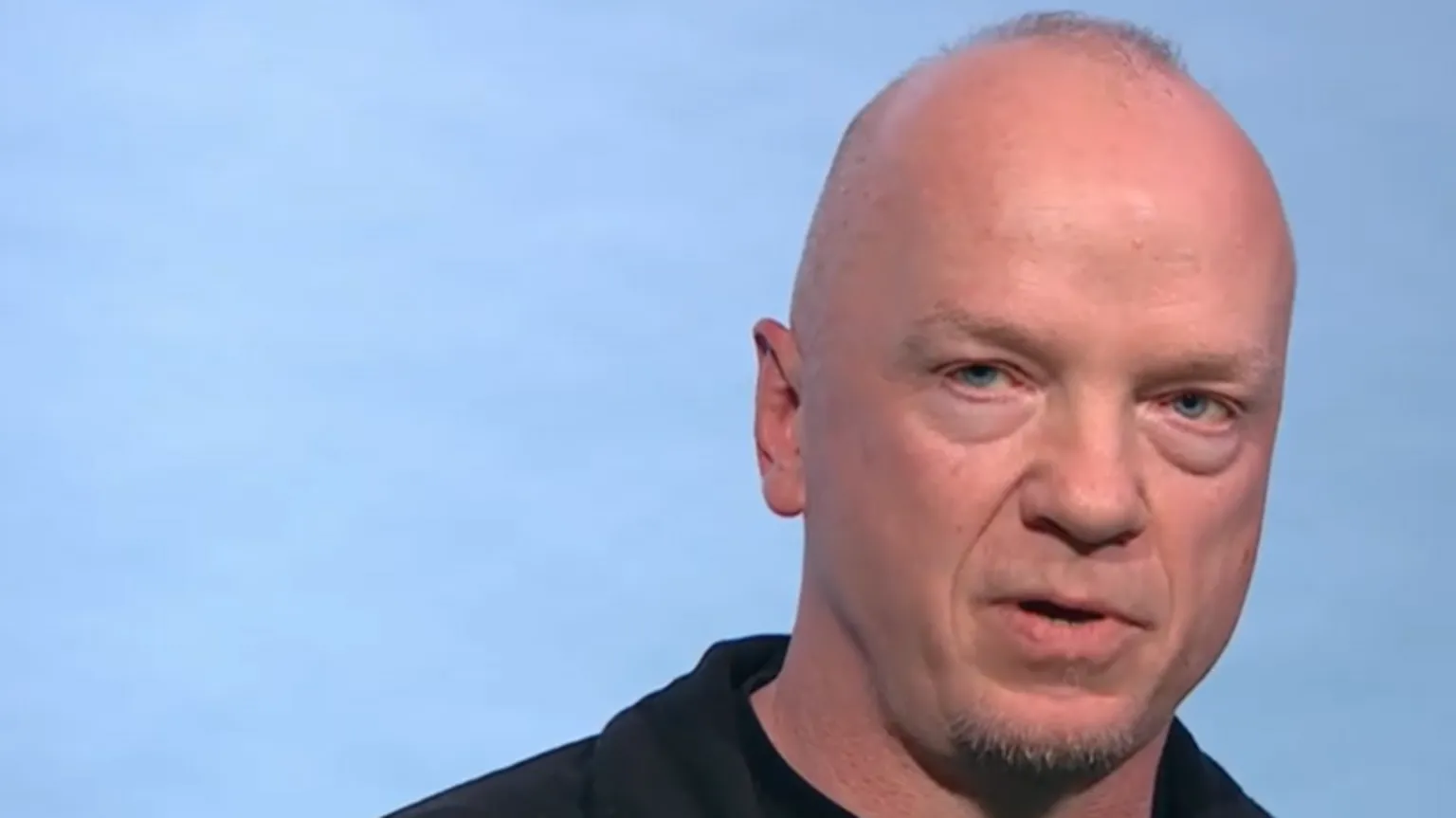 Fabulous A bald man with stubble, wearing a black shirt and looking at the camera with a serious expression. He is against a plain pale blue background. 