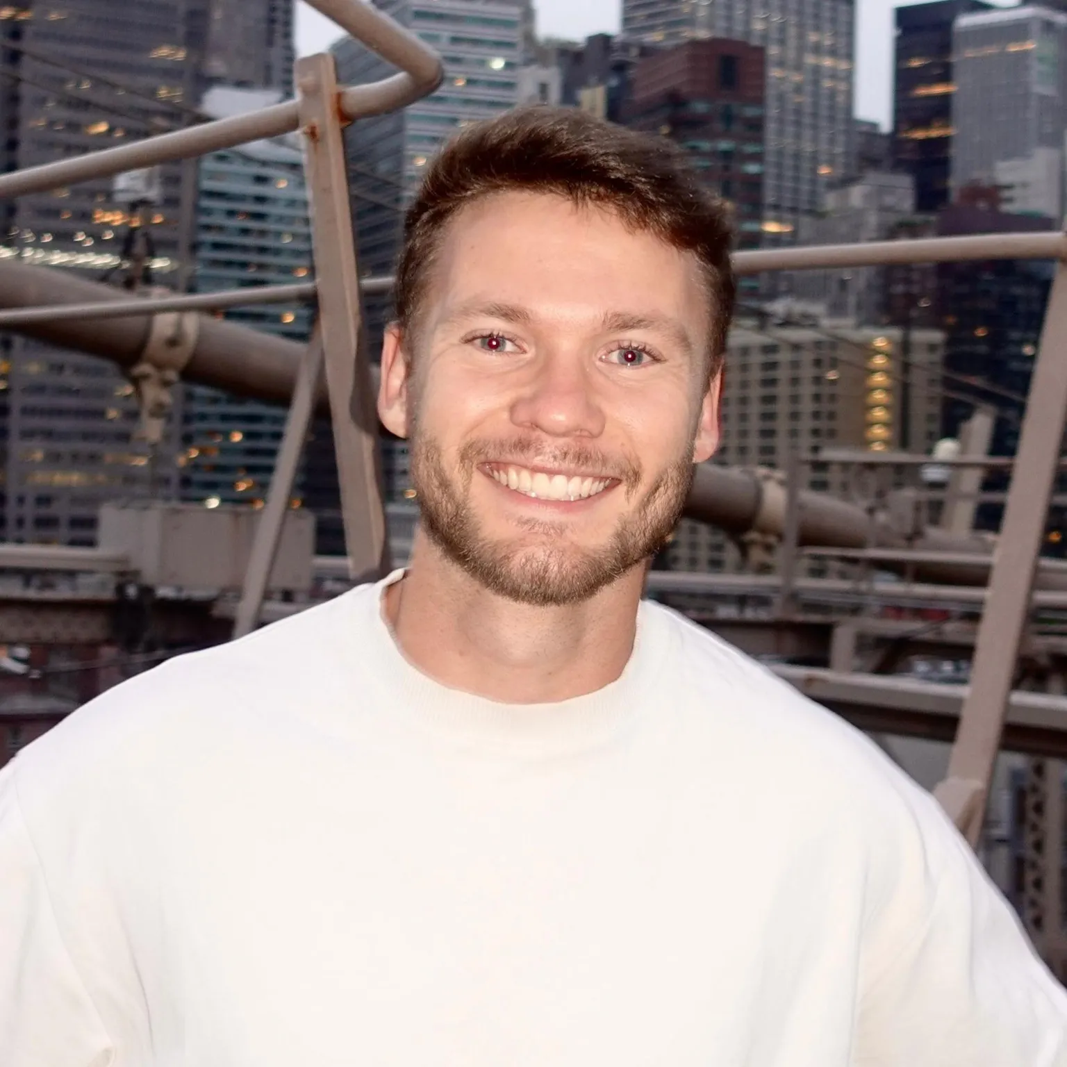Daniel Bayen A man with short blonde hair and a beard, wearing a white t shirt and smiling at the camera. He is stood on a bridge with city skyscrapers behind him.