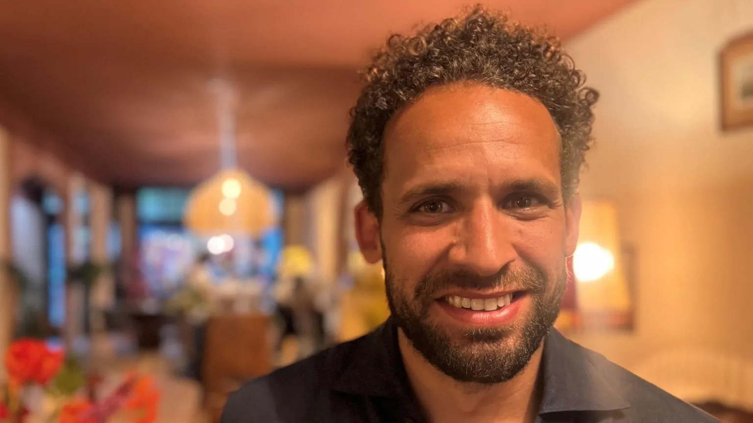 Close-up of a Alexander's face in the foreground, with short curly hair, a beard, and a dark buttoned shirt, positioned on the right side of the frame. In the blurred midground, a table displays a large arrangement of bright flowers in shades of orange and red. The background shows an indoor setting with warm lighting, brick walls, arched doorways and a hanging pendant lamp above a table further back. 