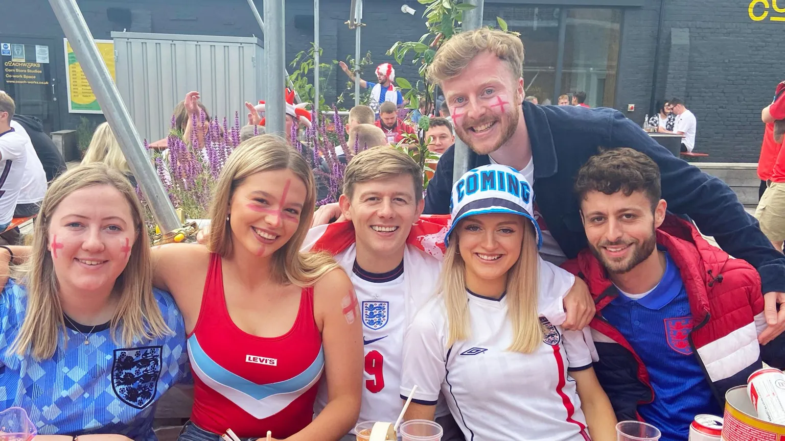 Sam O'Brien West Sam, with England flags painted on his cheeks, smiling into the camera as he poses with five of his friends, all wearing England football T-shirts, hats and face paint. They're sitting outside in a beer garden, with several alcoholic drinks on the able in front of them.