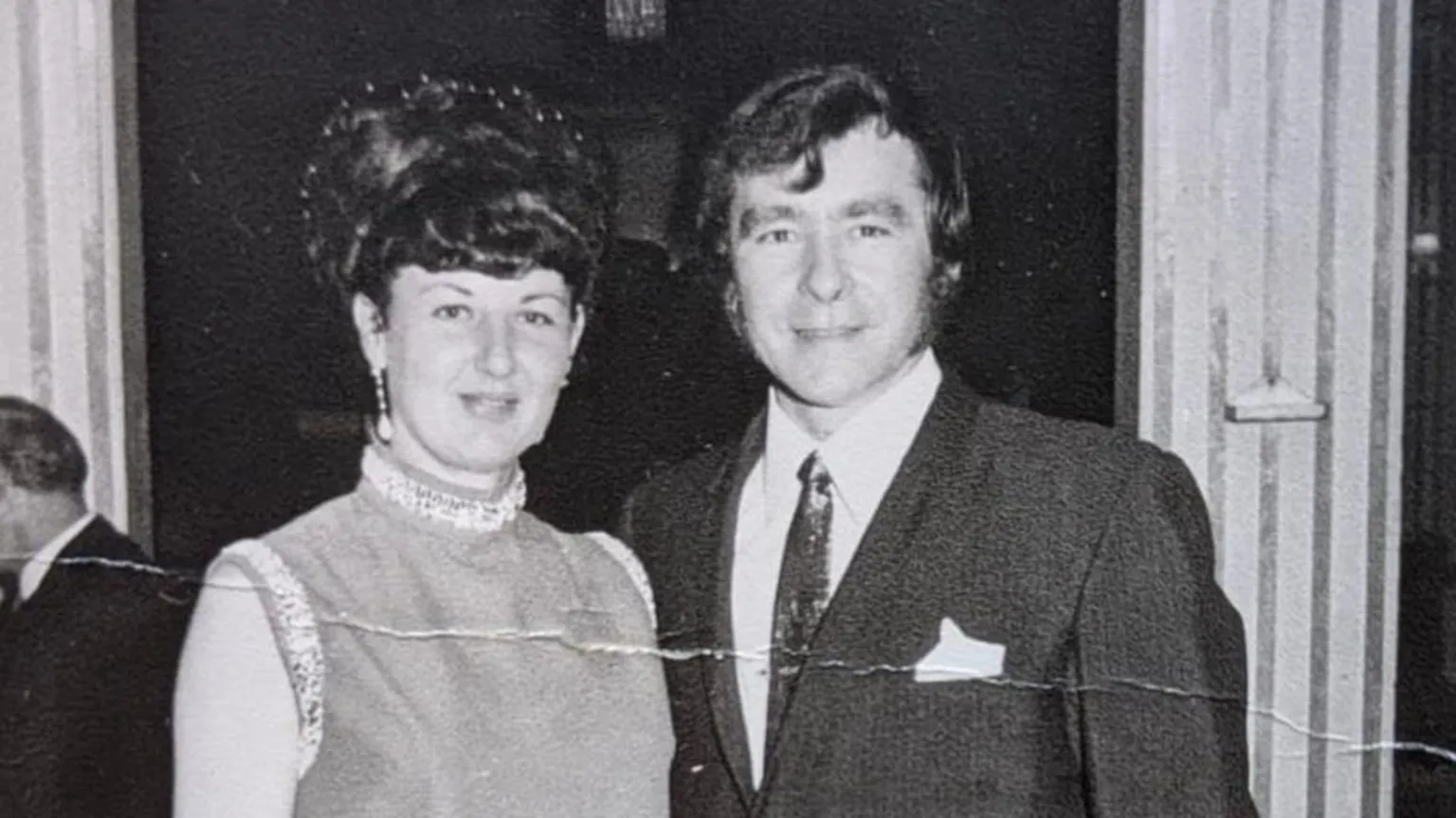 Ted Bush Ted is pictured in a black and white photo from the early sixties and has a quiff and sideburns and is wearing a dark suit with a tie and his wife Betty is wearing a cocktail dress with sequins around the arms for a black tie event.