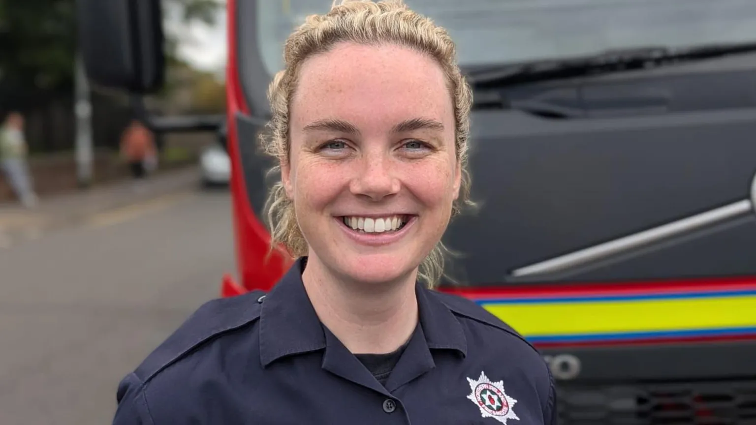 Ciara McGurk has curly blonde hair and is wearing a NIFRS navy uniform. She is smiling and standing in front of a fire engine.