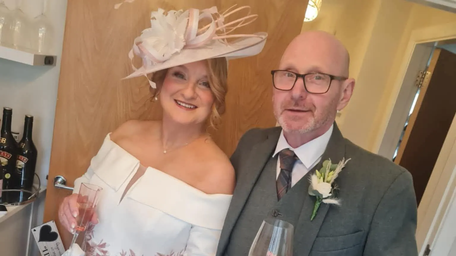 KAY DONALD Kay and Barry are a couple in their middle years. She is wearing a wedding outfit of cream hat and off-the-shoulder top and is holding a glass of champagne. He is in a tweed suit with a tartan tie and a heather button. He too is drinking champagne and they are beside a drinks cabinet.