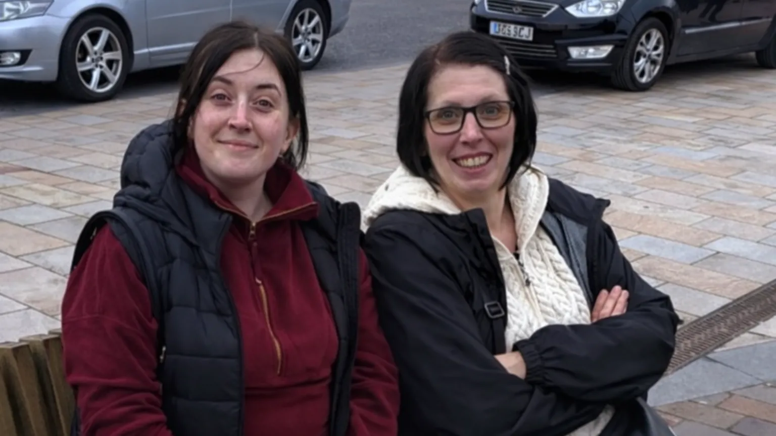 RACHEL SHANKLAND Two women who look very alike with dark hair, sit side by side on a bench, smiling. They are dressed for colder weather. There are cars behind them parked on the street.