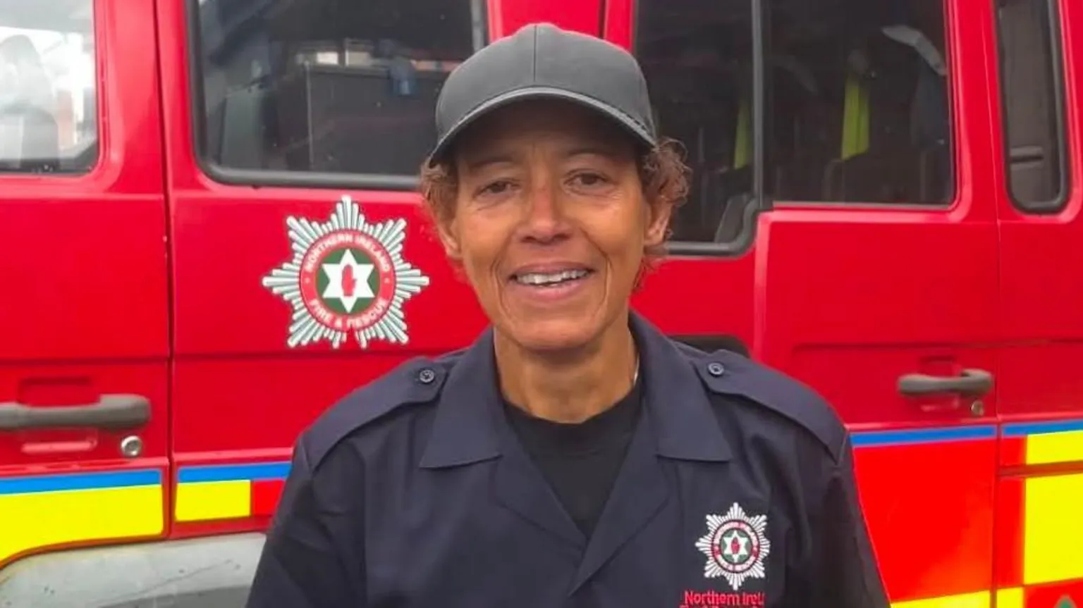 Lisa Osuere is wearing a navy NIFRS uniform, she is also wearing a black baseball cap. She has curly brown hair and is standing near a fire engine. 