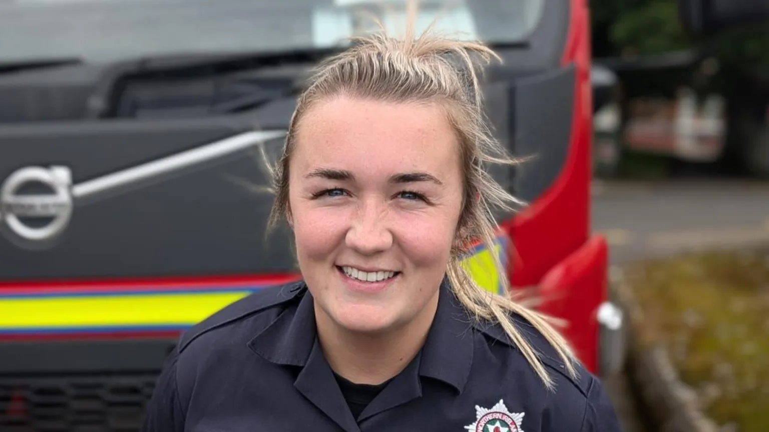 Michaela McGuigan has long blonde hair and is standing near a fire engine in her navy NIFRS uniform.