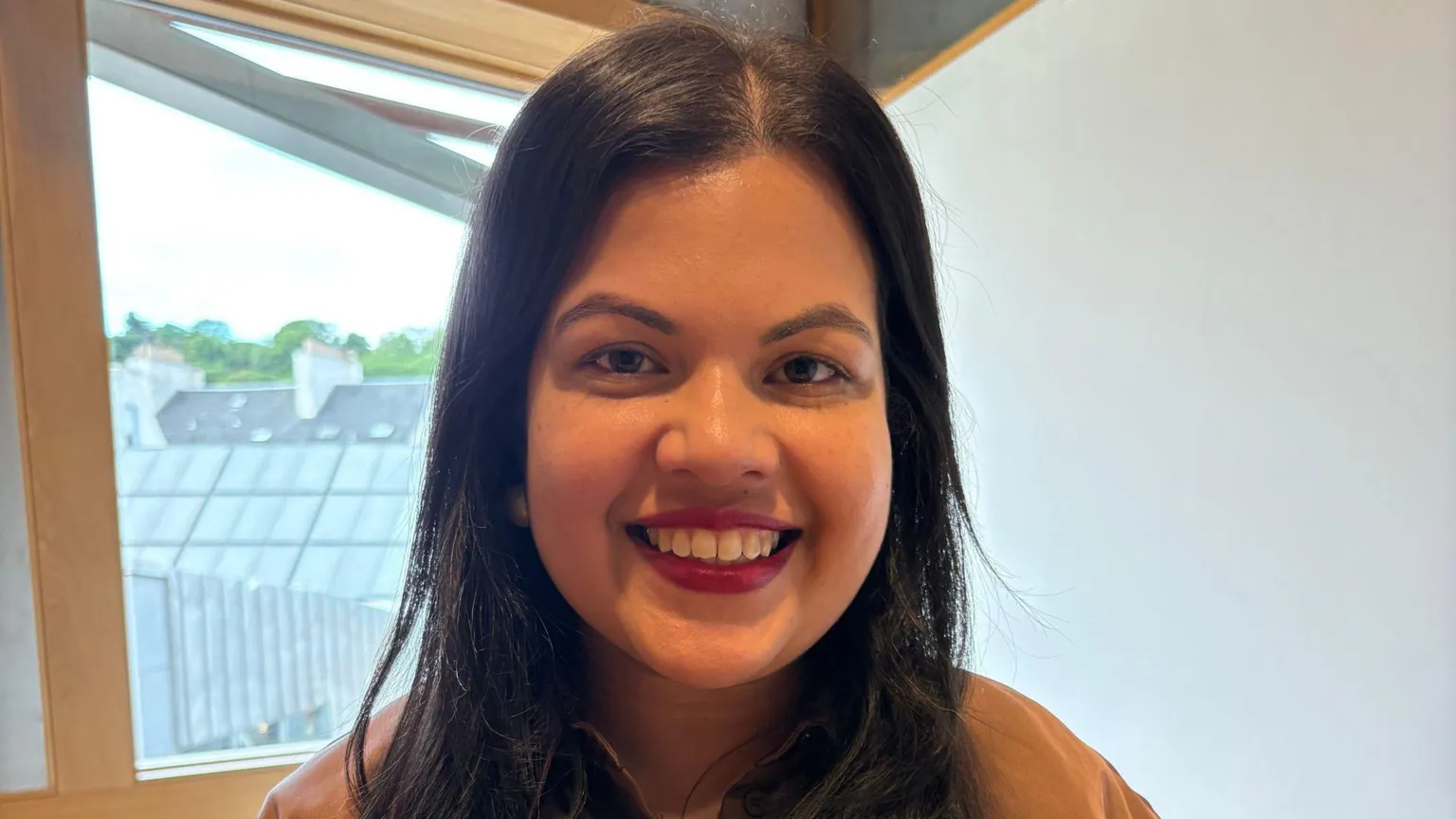 Simita Kumar has long dark hair and smiles at the camera. She wears a brown top and stands near a window