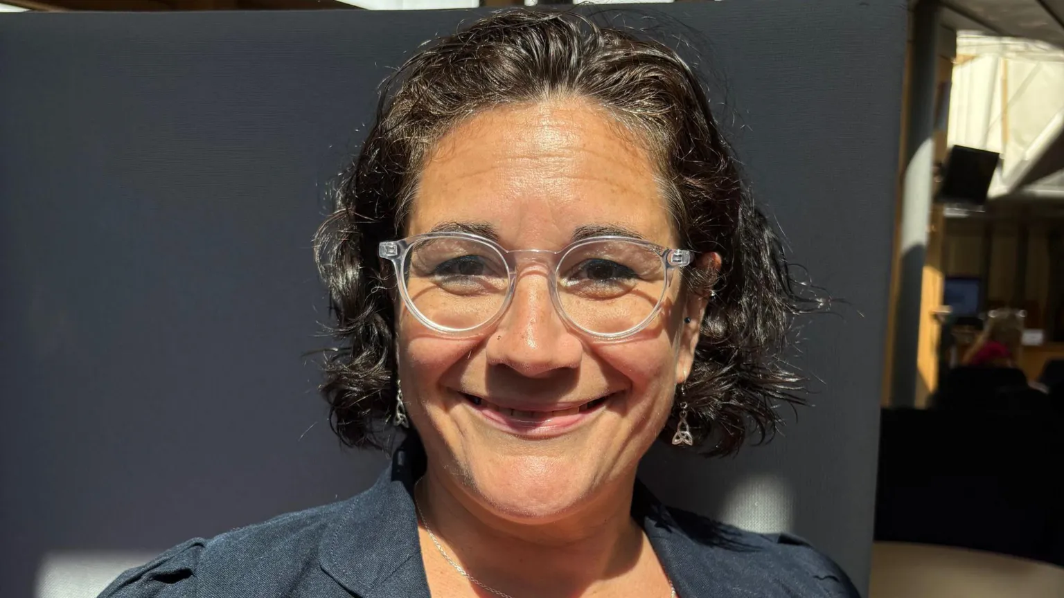 Katie Hagmann wears clear glasses and a dark blazer. She has short dark hair and smiles at the camera