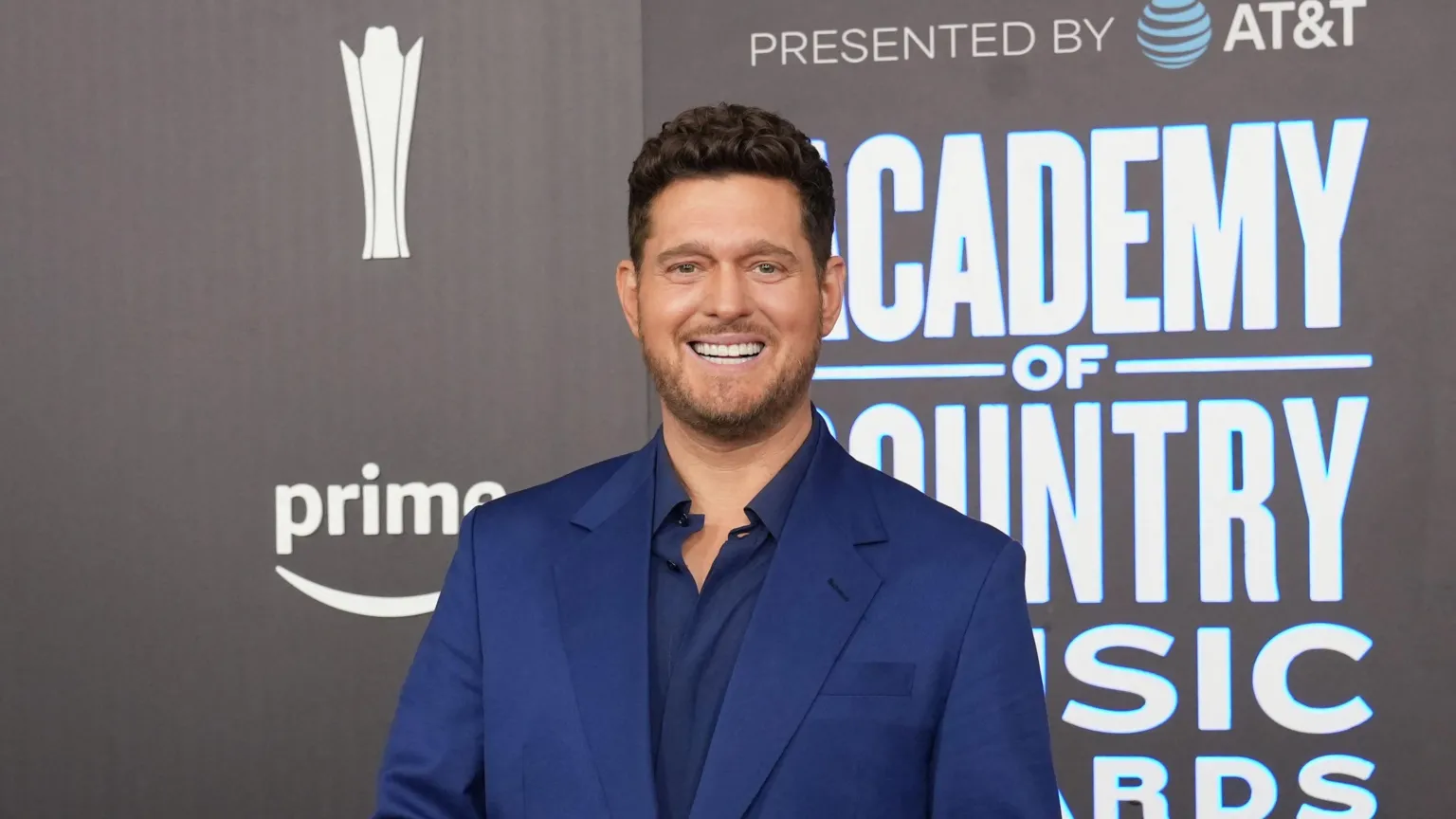 A man, Micheal Bublé, he is wearing a navy suit, with a navy shirt underneath. He is at a premiere, behind him is a media wall with an advertisement for Prime.
