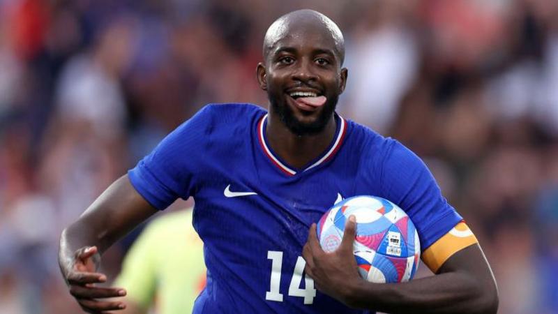 Mateta, in France's blue kit and with his tongue sticking out of his mouth, celebrates with the ball under his left arm after scoring against Spain in the 2024 Olympics men's football final
