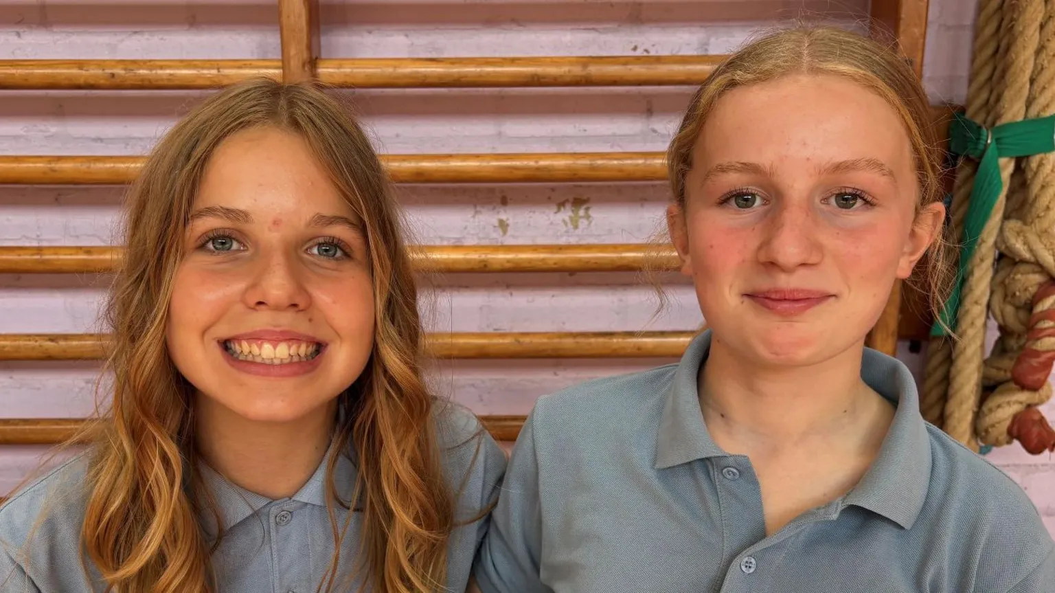 Myfi and Elin sit next to each other smiling at camera. Myfi, on the left, has curly blonde hair. Elin has blonde hair in a low ponytail. Both wears light blue uniform polo tops. 