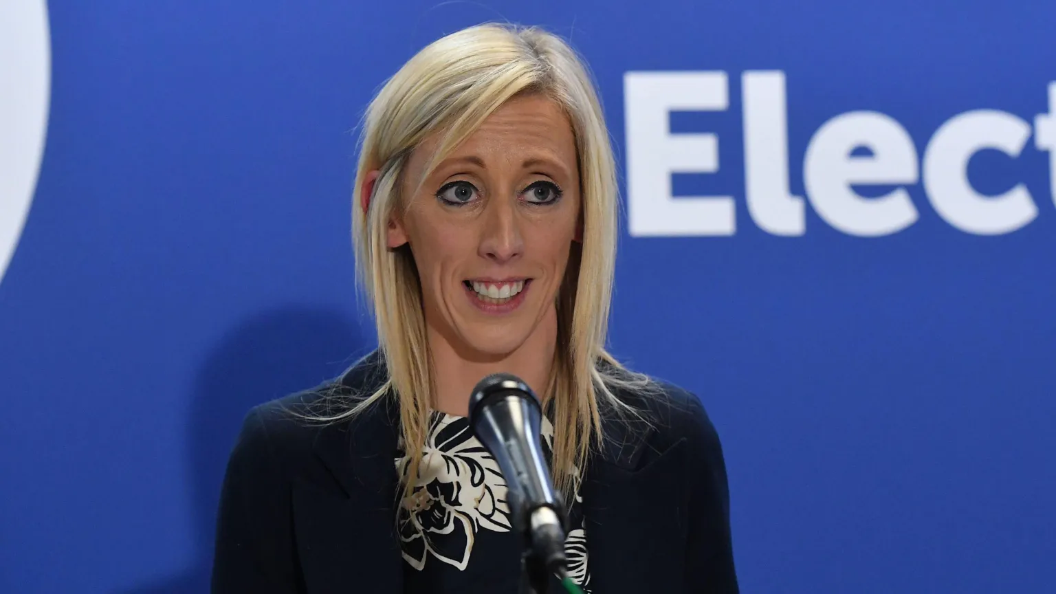 Carla Lockhart, with long blonde hair, wearing a black and white top and a black blazer. She is standing in front of a blue wall.