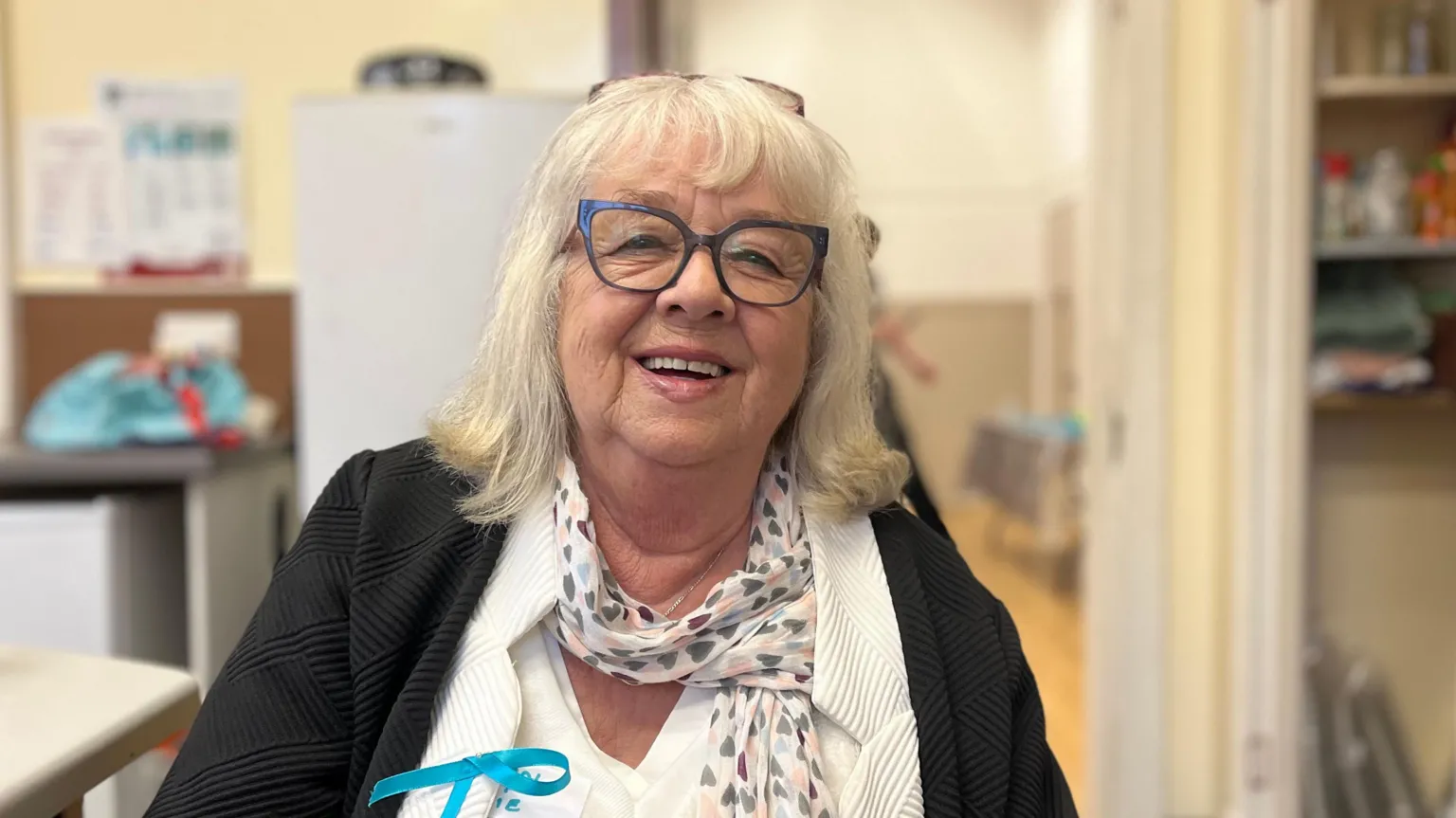 A white haired woman with glasses, wearing a black jacket and white top, smiles at the camera