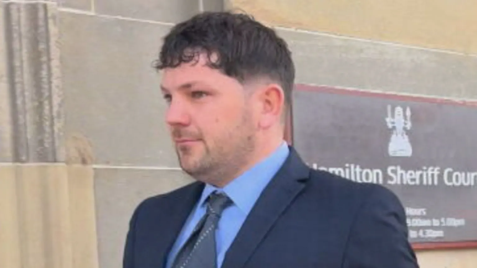 Tom Nicholson Jr has short brown hair and wears a dark navy suit with a blue shirt and grey tie outside Hamilton Sheriff Court