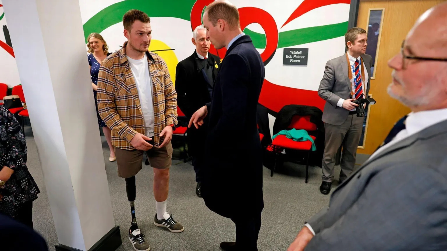 Wales Air Ambulance Richard and Prince Williams stand facing each other talking. Richard is wearing shorts showing his prosthetic leg. He has short, dark hair and is wearing an open button down check shirt over a white t-shirt. Prince William is wearing a dark suit and overcoat.