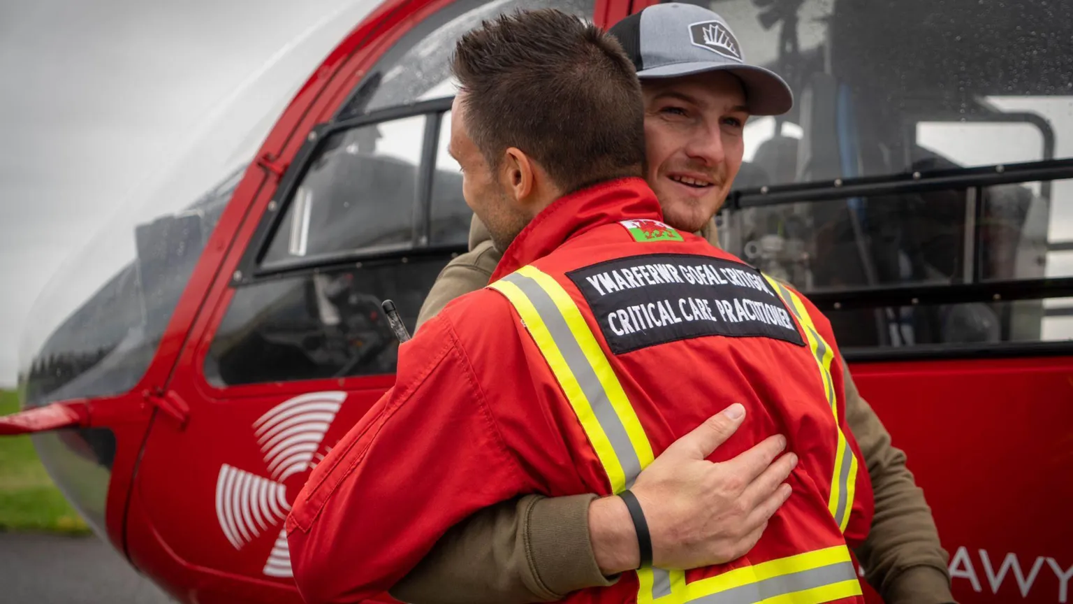 Wales Air Ambulance Two men embrace in front of a red Wales Air Ambulance helicopter. The man facing away from the camera wears medics jacket that says critical care practitioner. The other man, Richard Jones can bee seen with his face over the other man's shoulder, he is smiling, has light stubble and wears a grey baseball cap.