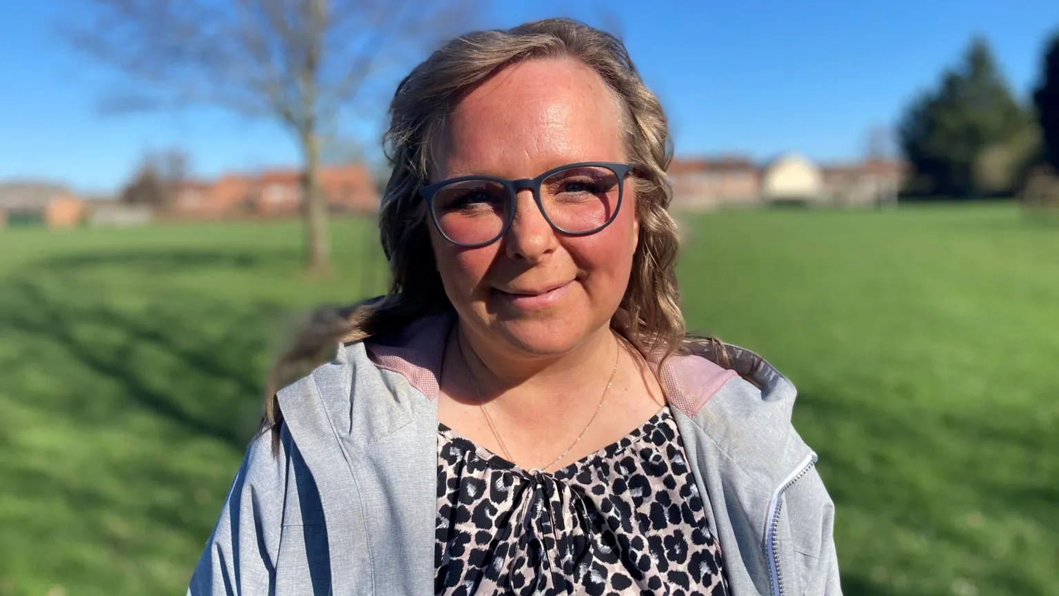 Karen Bentham is outdoors with a tree and grass in the background. She has shoulder length hair and is wearing a leopard print top with a grey coat over it.