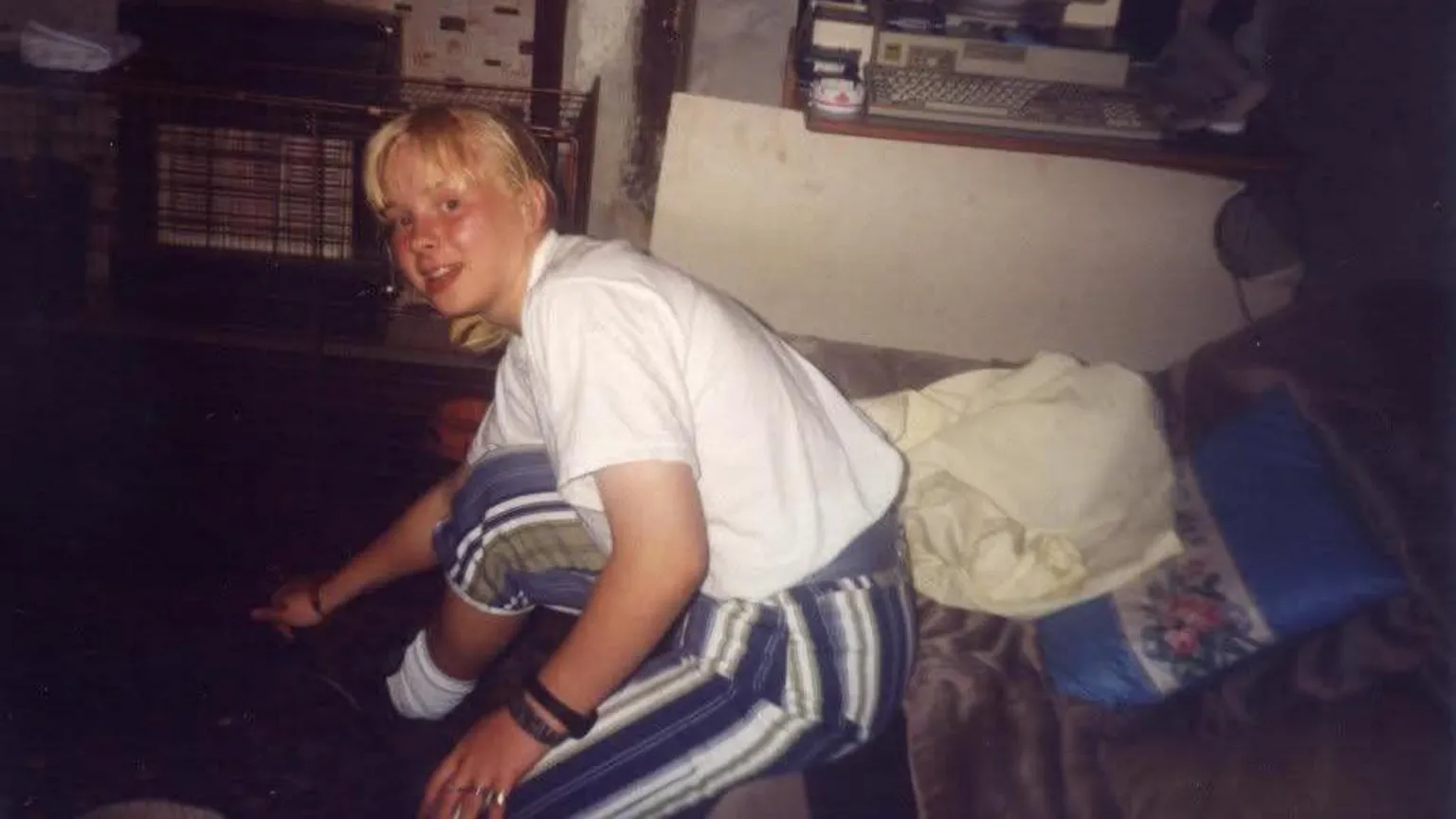 Karen Bentham is pictured as a child of 14. She is wearing blue and white striped trousers and a white shirt. She has blond hair and is perched on the edge of a seat.
