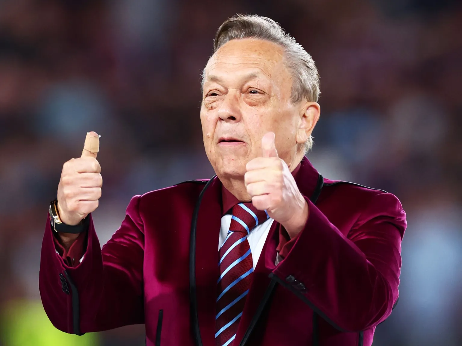 Man in a claret-coloured blazer, and claret and blue tie, stands with both his thumbs pointing up. The background is blurred.