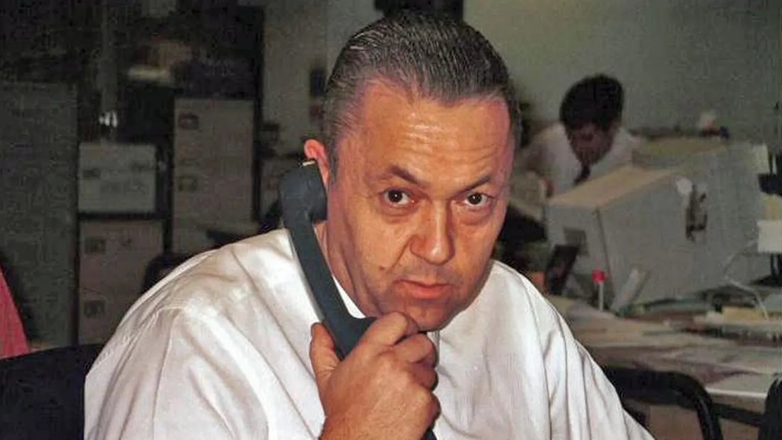 A middle-aged man looks ahead at the camera. He is wearing a white shirt and is holding a telephone receiver to his right ear.