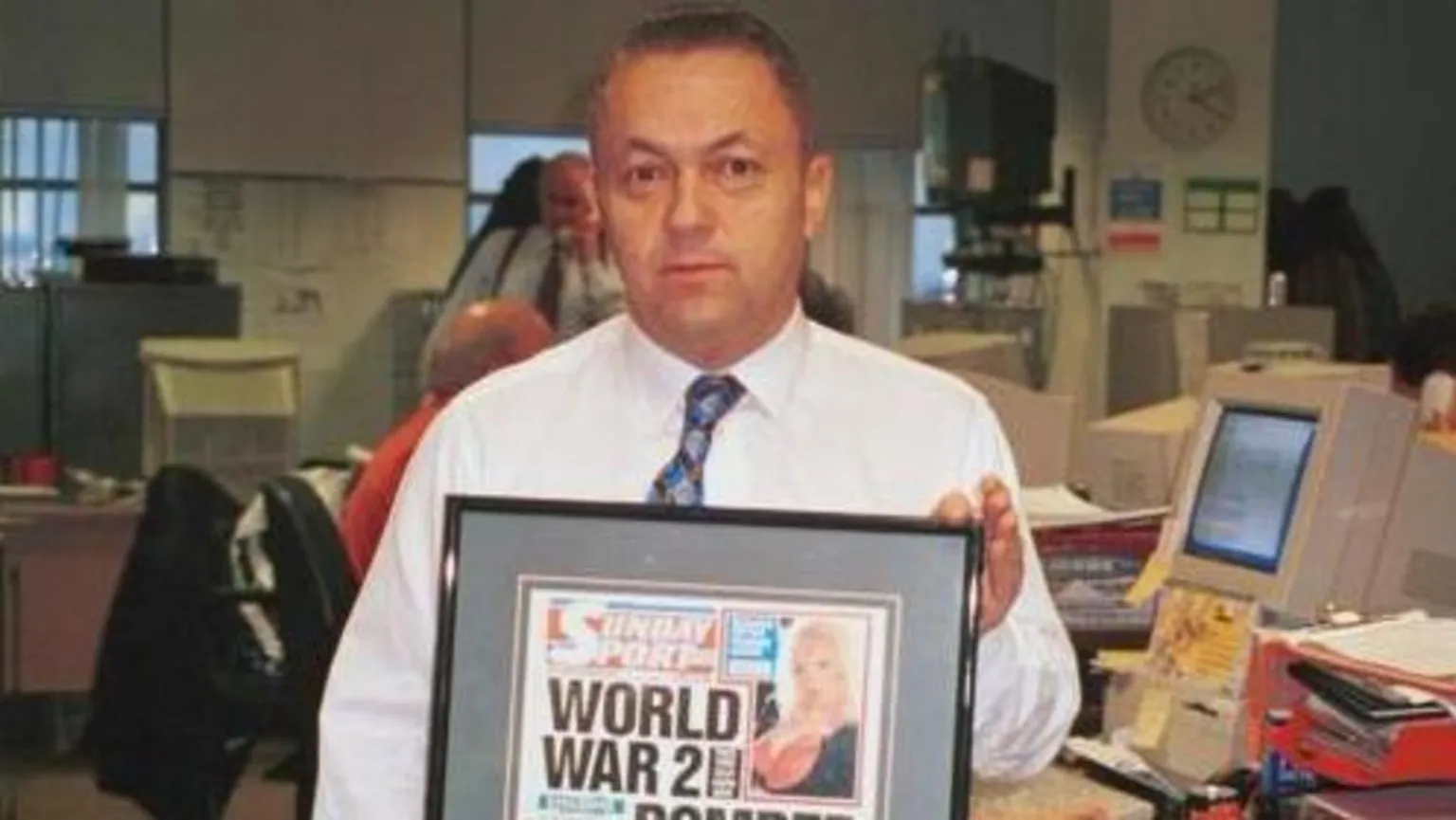 David Sullivan, pictured in 1999 for an episode of the BBC Two show Back to the Floor, in shirtsleeves with a tie, holding up a framed Sunday Sport front page with a blonde model in the top right corner and the headline: