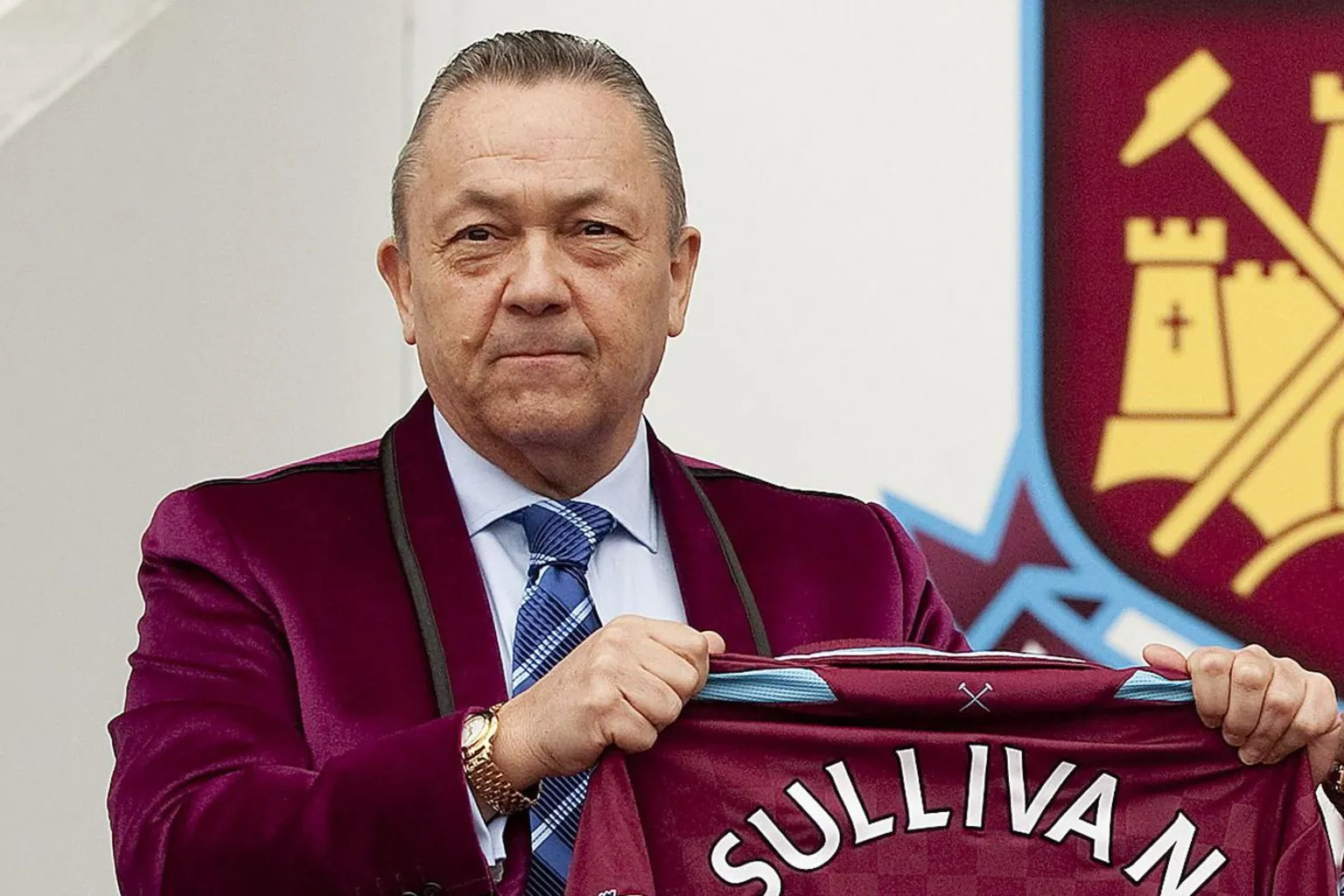 David Sullivan, wearing a velvet jacket in West Ham's claret, holding a club shirt with his name written on the back, while standing in front of the club crest.