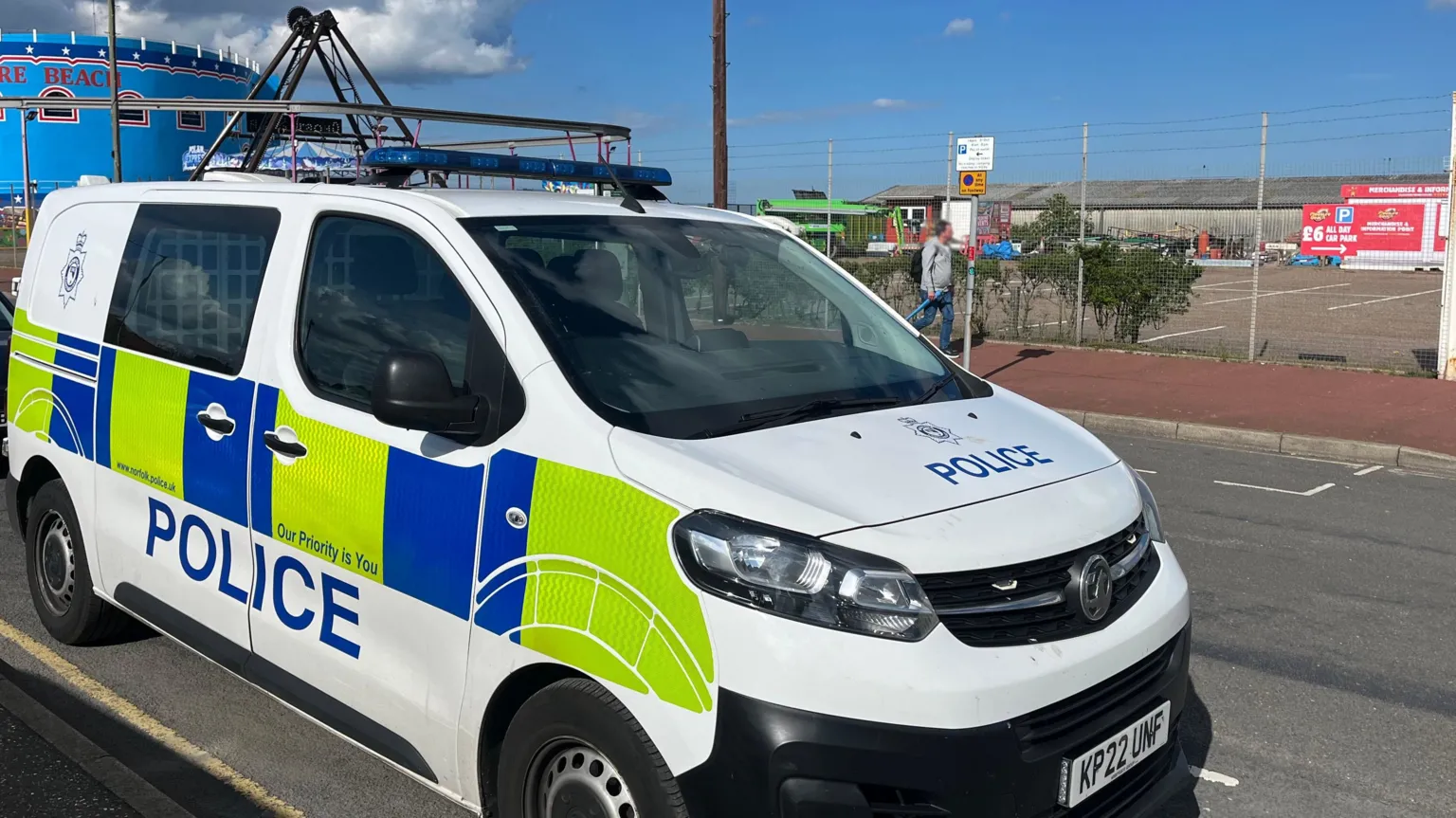 Qays Najm/BBC A marked police van is parked on a road next to a beachfront. There are fairground rides in the background behind a fence, and an individual is walking along a pavement.