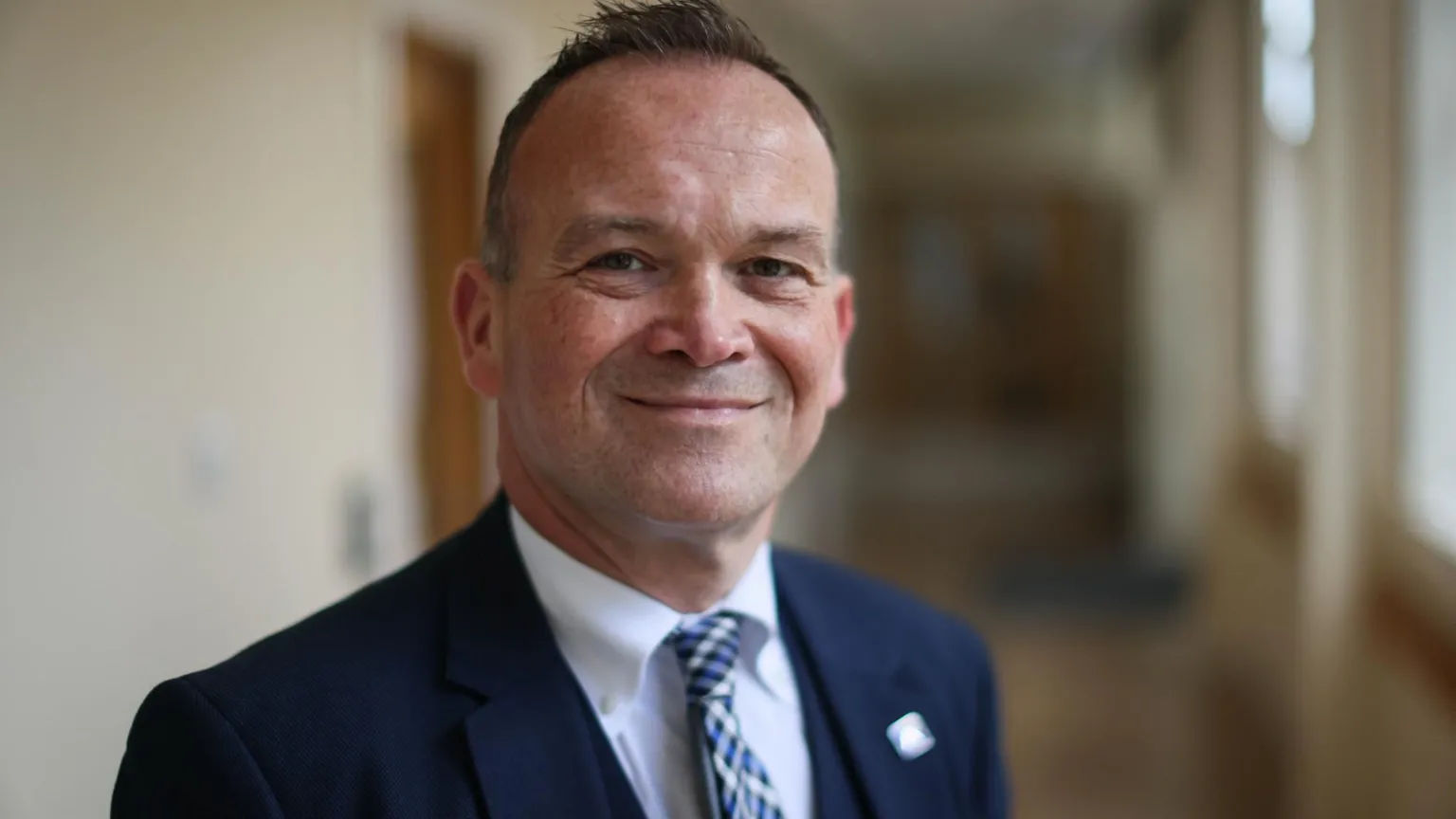 Laurence Findlay, a man with short, brown hair, standing in a school corridor. He is wearing a blue suit, white shirt and patterned tie.