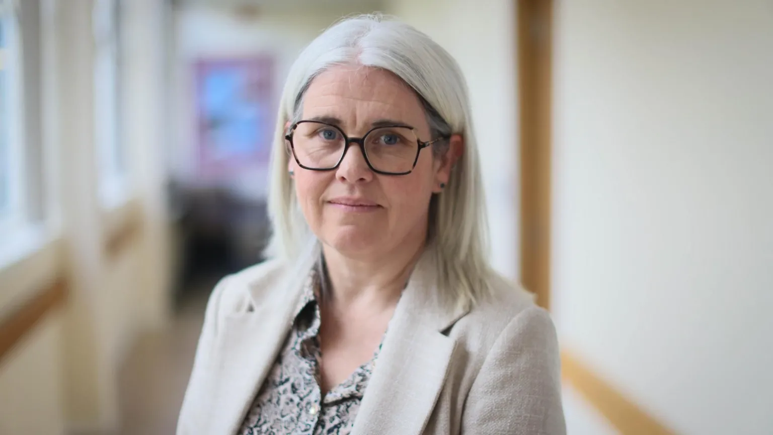 Louise Moir standing in a school corridor. She has shoulder-length, grey hair and glasses, and is wearing a cream jacket and a patterned top.