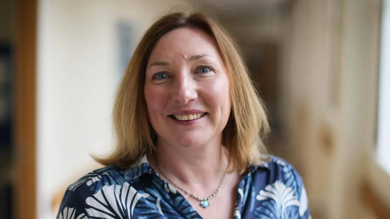 Katrina Suèr standing in a school corridor. She has shoulder-length, brown hair and is wearing a blue and white patterned top.