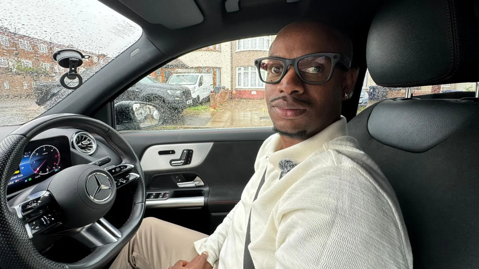 Donovan Smith, a driving instructor in south London sits at the sterring wheel of his car. He has shaved hair and wears glasses, wearing a cream summer jumper and beige trousers.