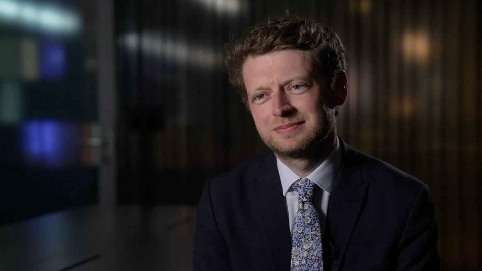 Connor Rand smiles, wearing a black suit, white shirt and floral tie, as he is interviewed in an office room