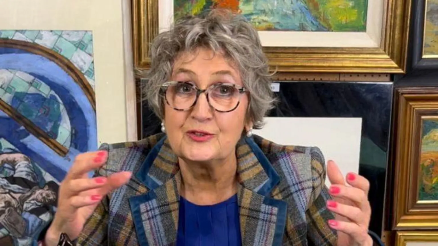 Great Western Auctions Anita Manning - a woman with glasses and grey hair, has her hands raised in the air. There are various paintings and antiques behind her.