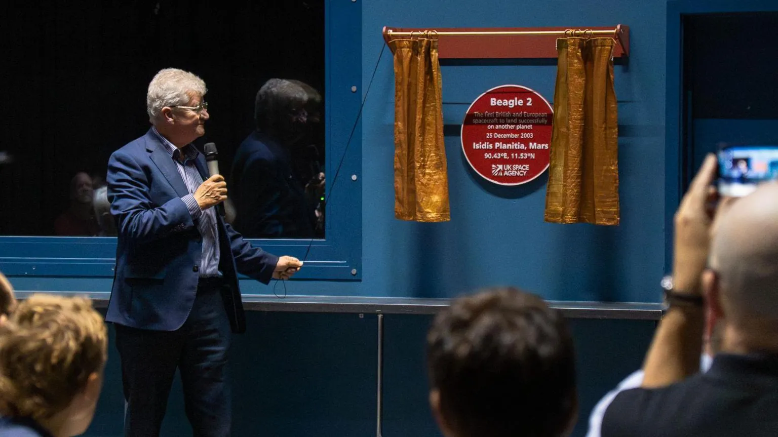 Department for Science, Innovation and Technology A red plaque is on a blue wall, revealed by a man with grey hair pulling on a rope that opened some gold curtains