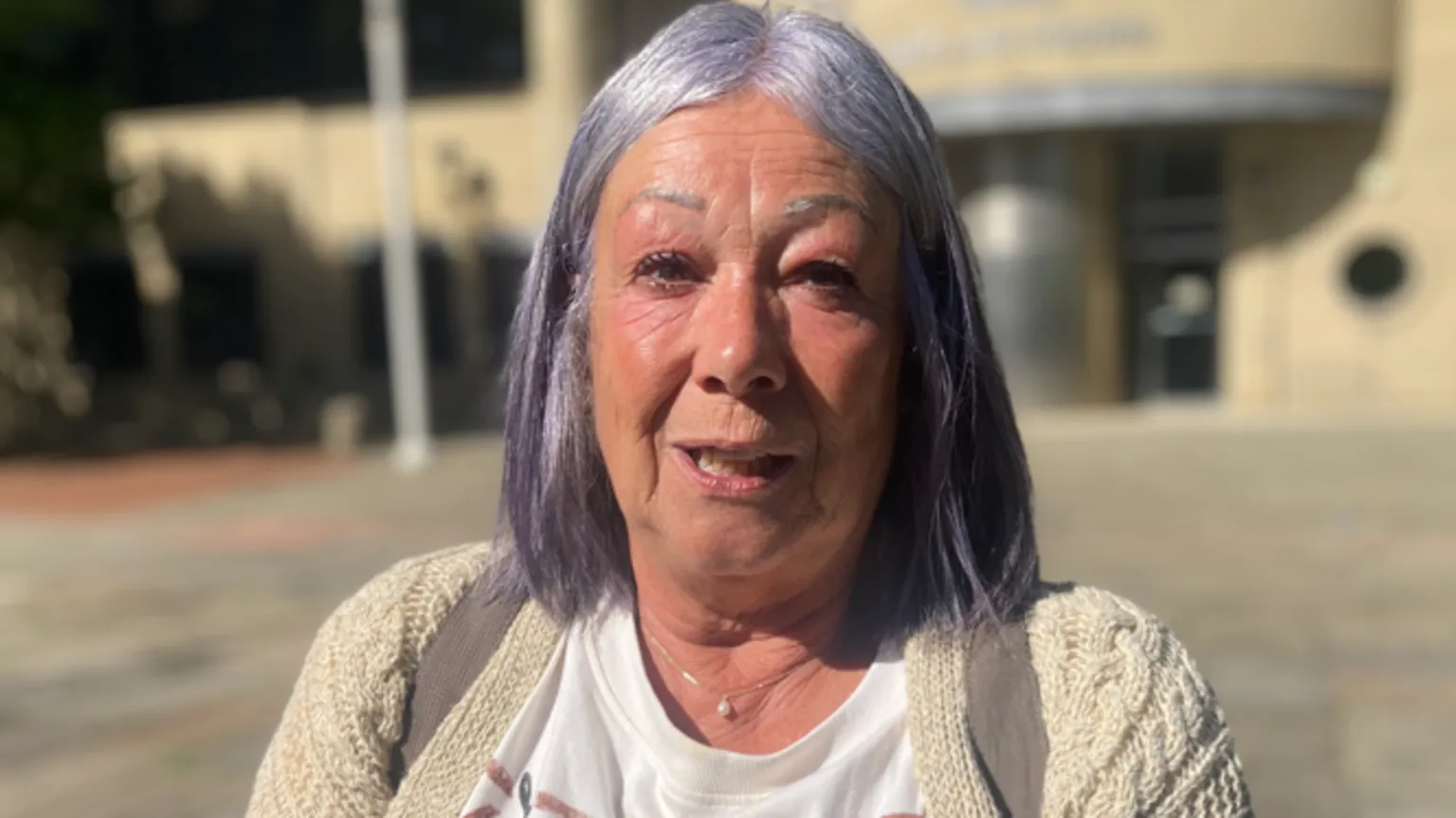 A woman with silvery purple hair looks directly at the camera outside a court building.