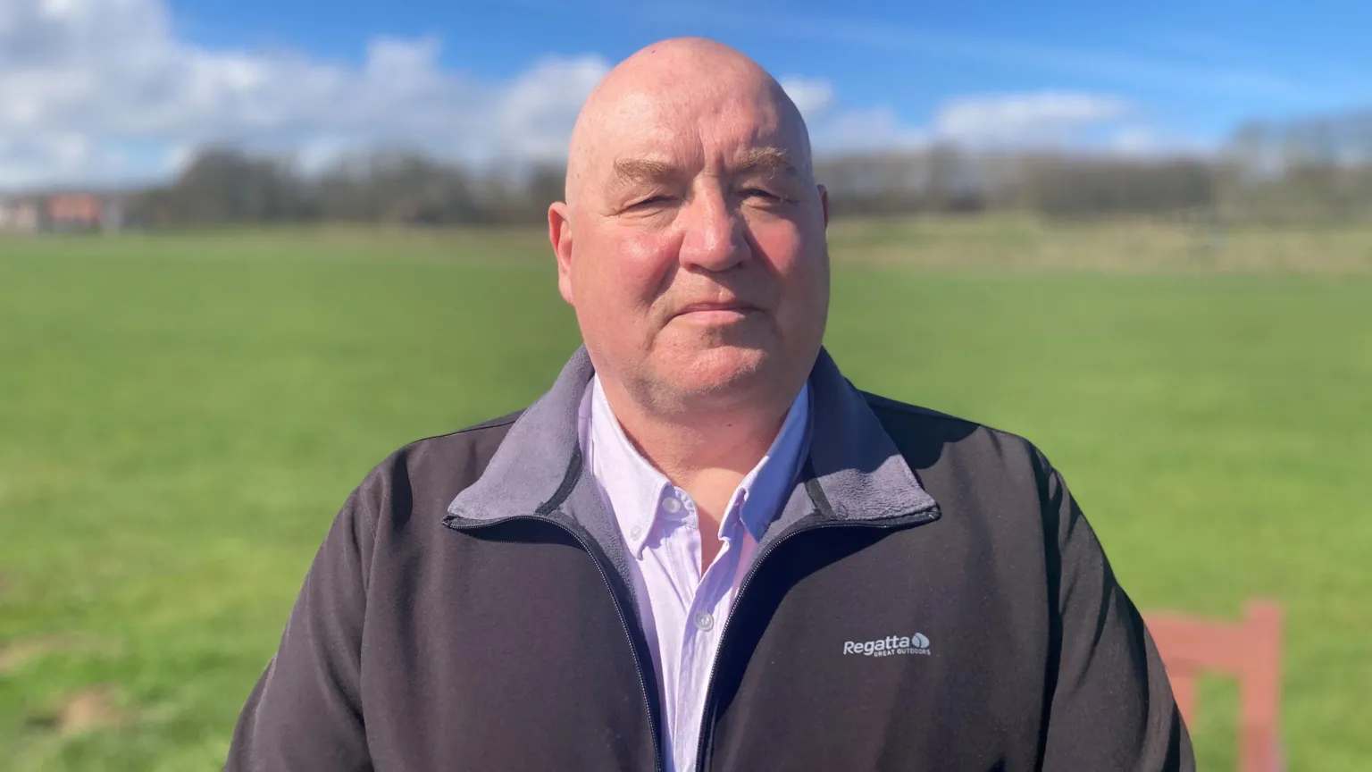 A bald man stands in a shirt and fleece against a rural backdrop