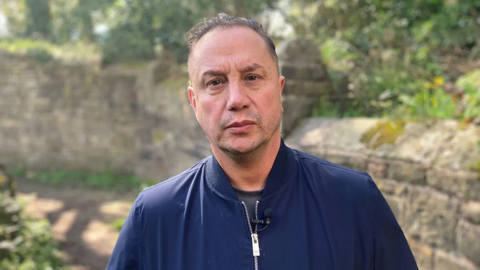 A man with short dark hair wears a blue jacket and stares at the camera