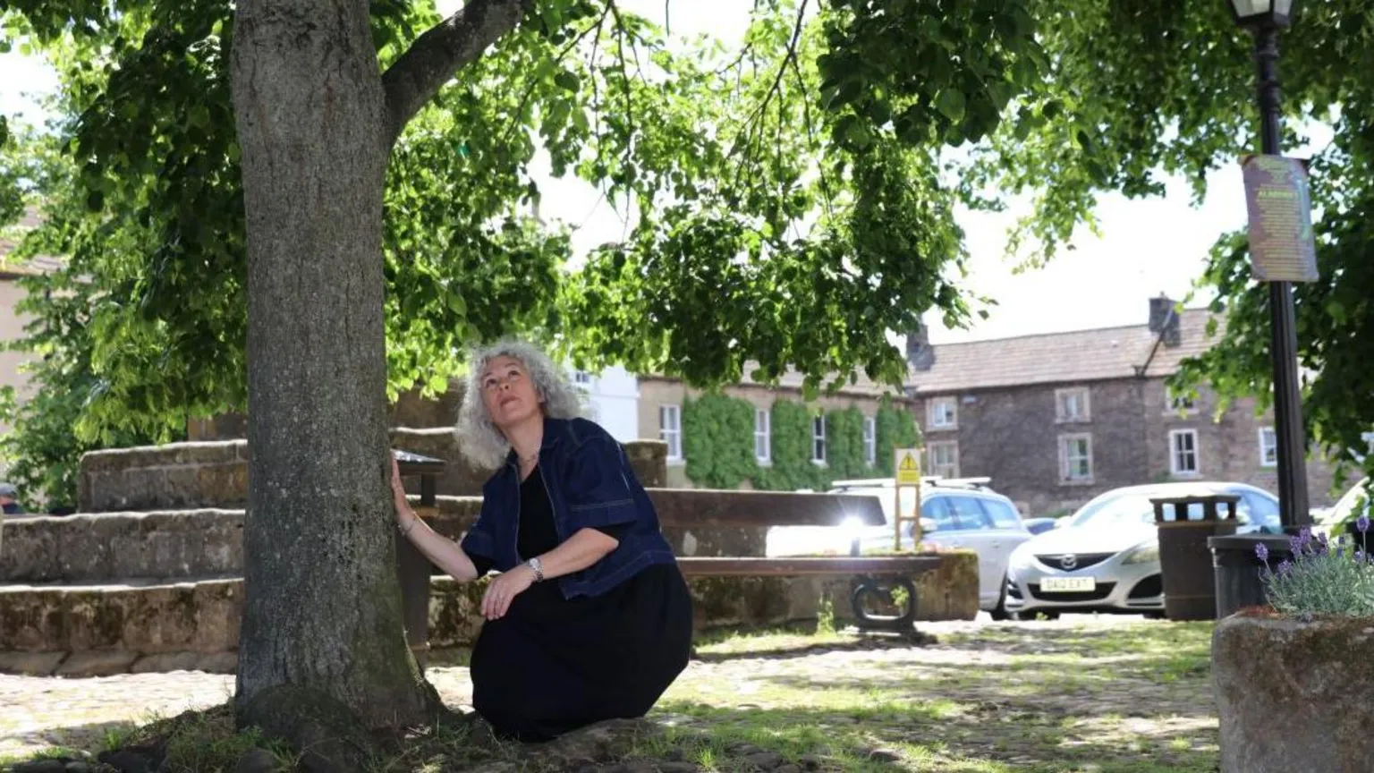 North Yorkshire Council A village market place on a bright, sunny day, with stone buildings and parked cars. In the foreground, a woman is crouching beside the base of a tree. One hand is placed near the tree trunk, while the other hand rests lower down, close to the ground.