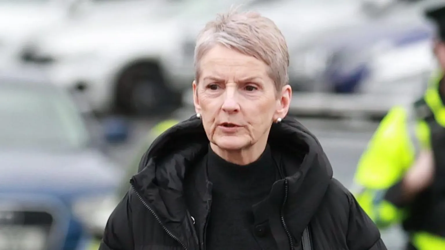  A woman with short grey hair walking through a carpark wearing a black coat and black high necked top.