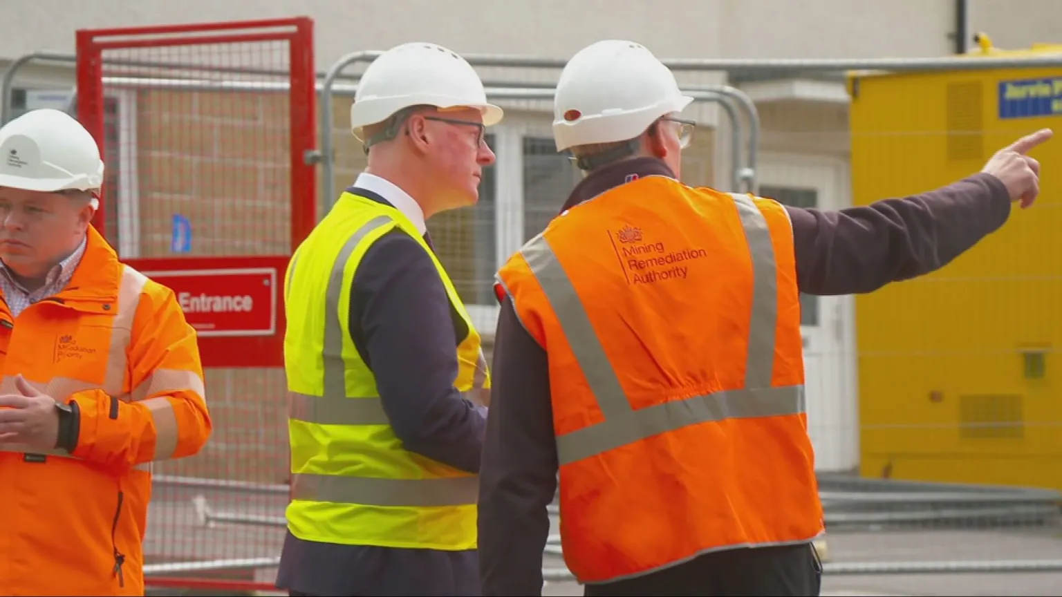 A man in orange hi vis points to something as John Swinney, wearing yellow hi vis, looks on