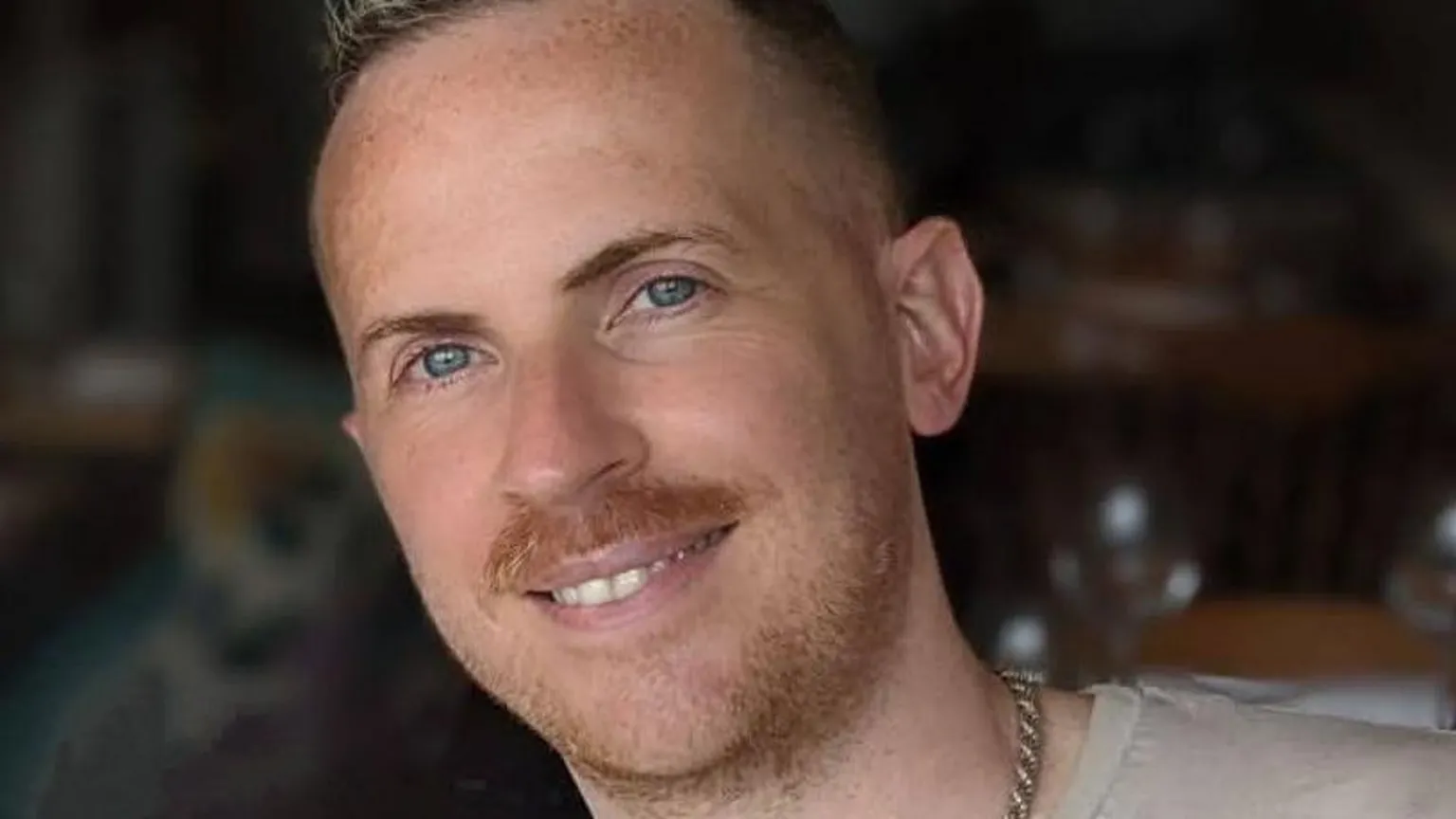 Steven Elliott is smiling at the camera. He has a moustache and a light beard and is wearing a thick chain around his neck