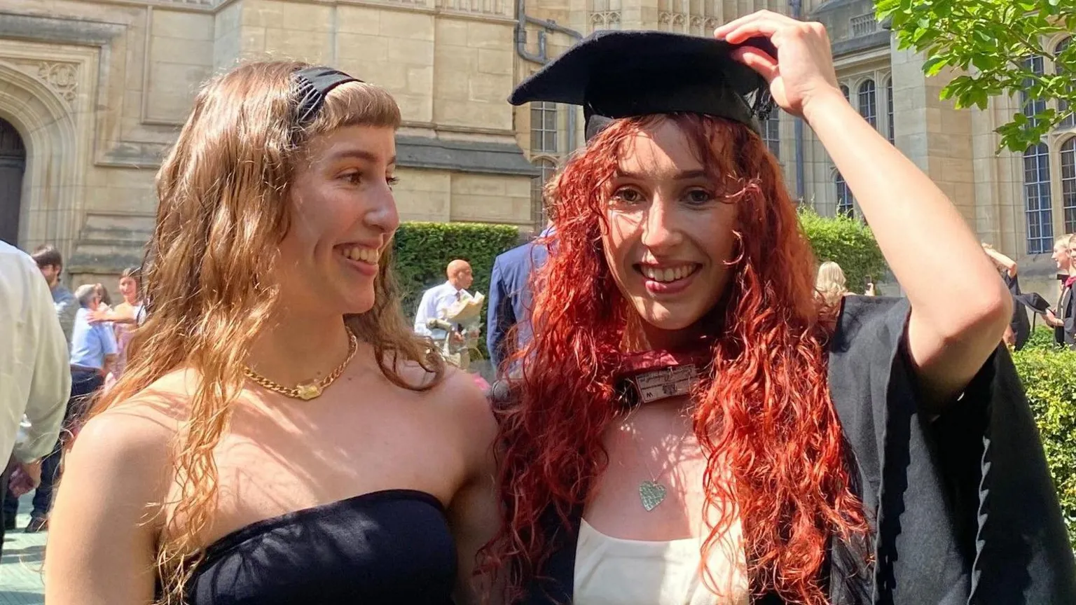 Grace and Caitlin stand in front of a university building together. Grace has long blonde hair and wears a black dress. Caitlin has died red hair and is wearing a graduation hat and gown.