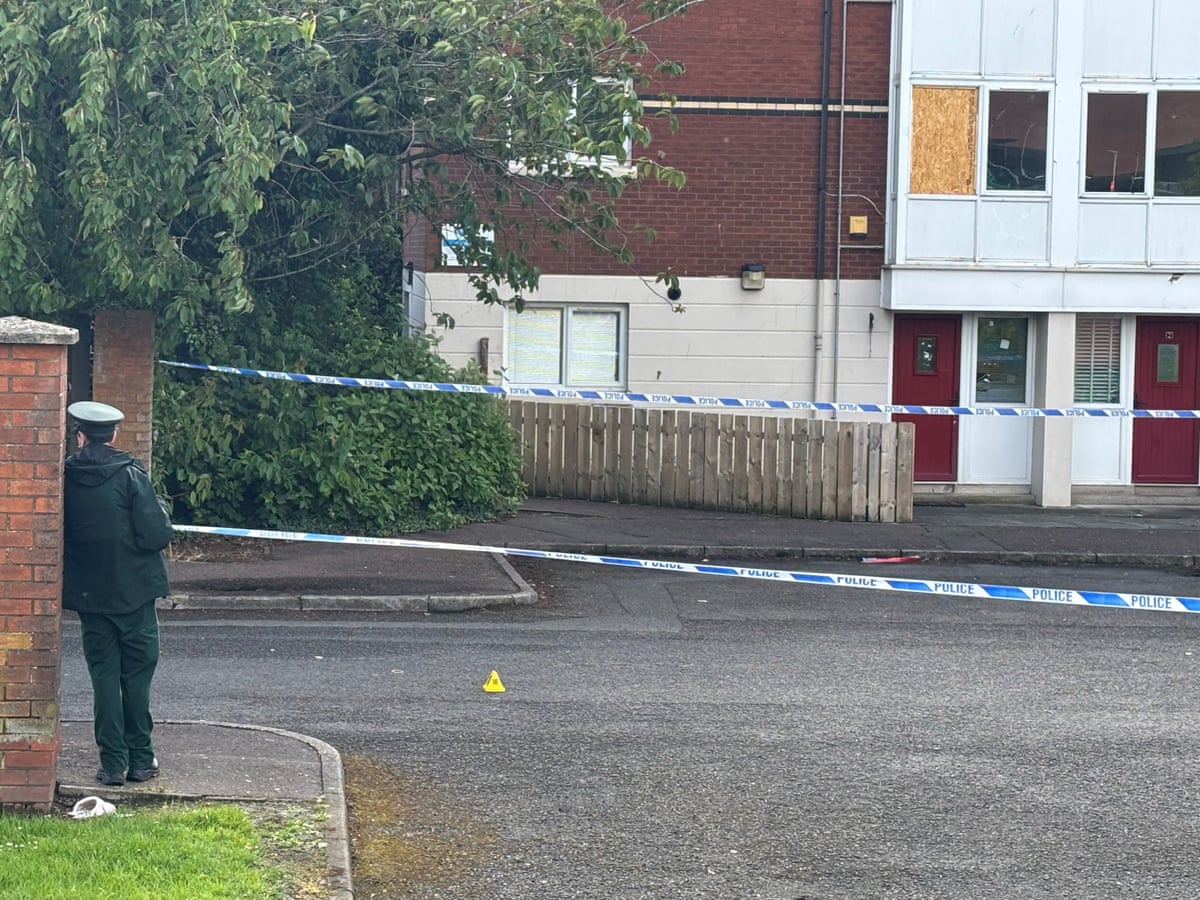 A police officer stands at the edge of the cordon, where police tape stretches across the road in front of apartments. One apartment window is boarded up