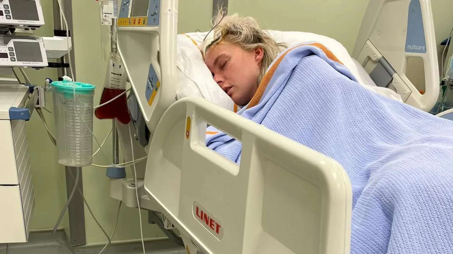 Mererid Rees Mererid is in a hospital bed with a blue blanket covering her and a white dressing on her head after surgery.