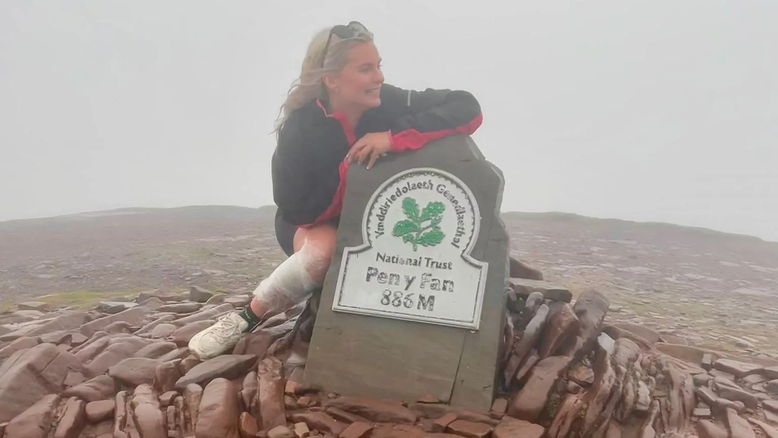 Mererid Rees A woman crouched in front of the Pen-y-Fan sign on the top of the summit. She's wearing shorts, a tacked and trainers with a white bandage on her right leg. She's surrounded by brown slab stones.